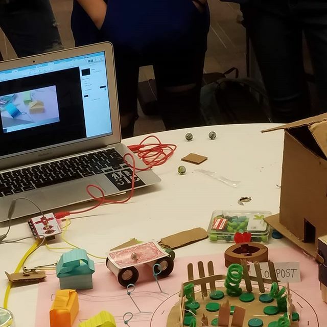 In the foreground, a model neighborhood made of paper, popsicle sticks, and plastic pieces attached by wires to a laptop in the background.